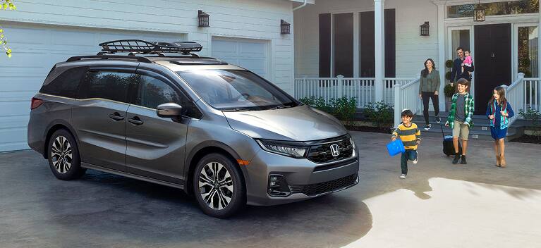 2027 Honda Odyssey Elite in Modern Steel Metallic front parked in a residential driveway with a family (Minivan).