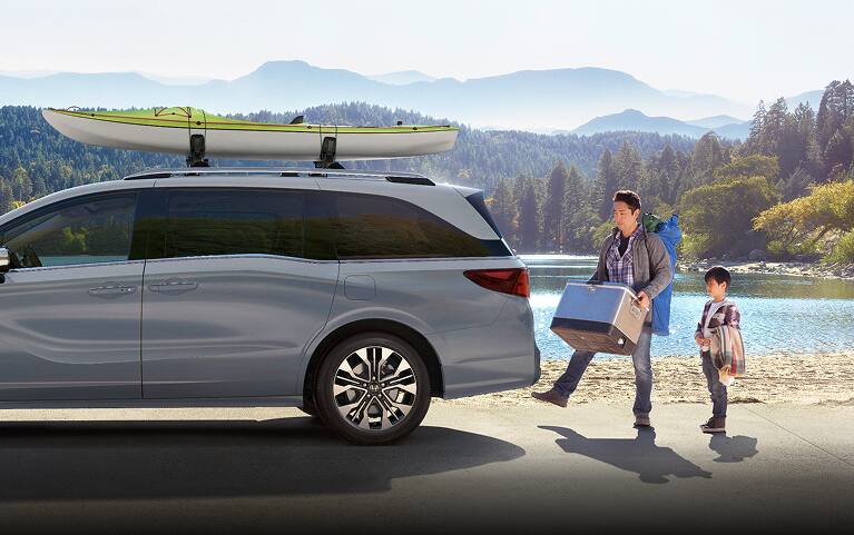 2027 Honda Odyssey Elite in Sonic Gray Pearl profile parked by a scenic lake with a family loading gear (Minivan). 2027 Honda Odyssey Elite in Sonic Gray Pearl profile parked by a scenic lake with a family loading gear (Minivan).